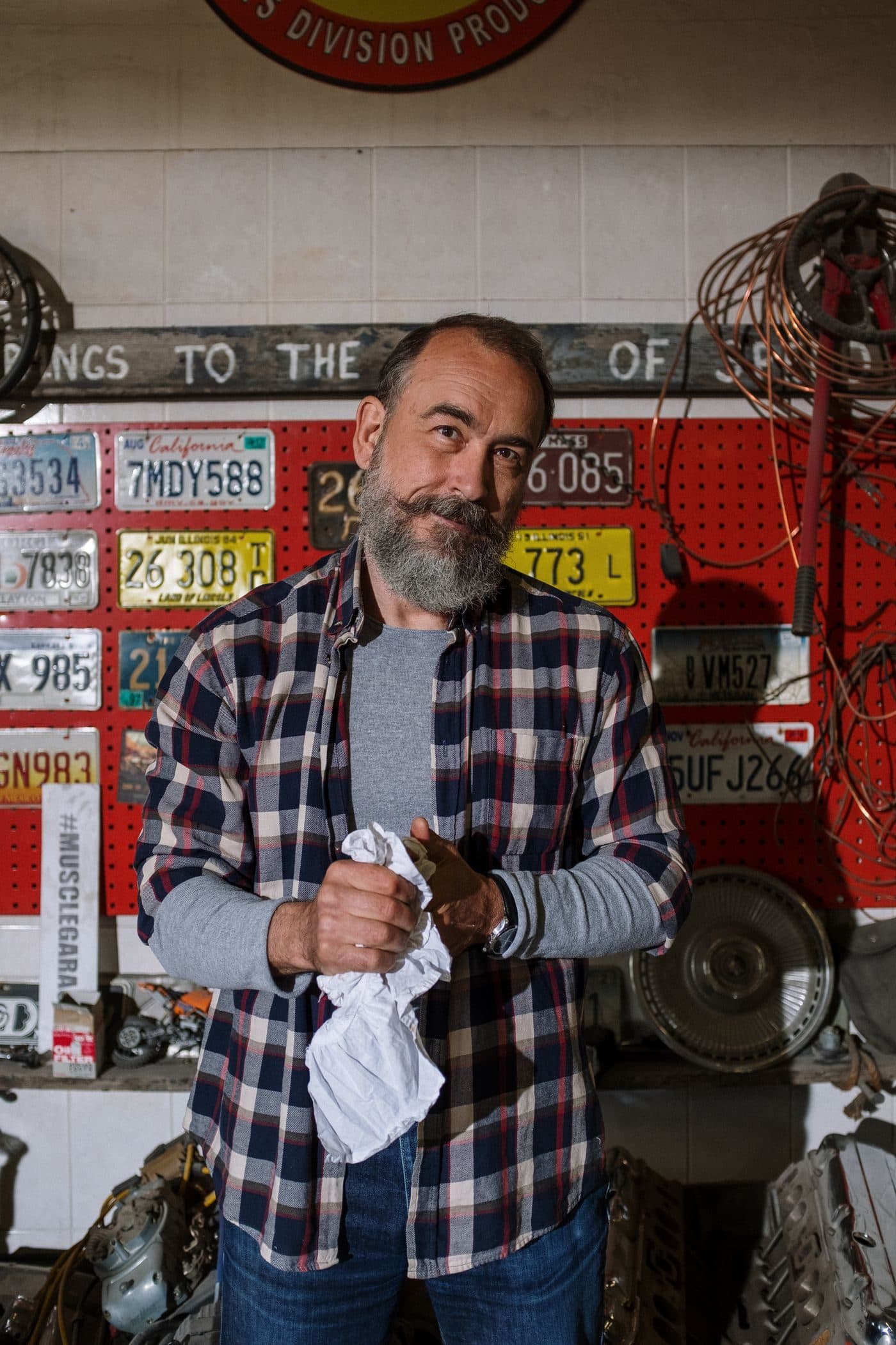 An auto shop owner in his garage, in front of a wall of old license plates
